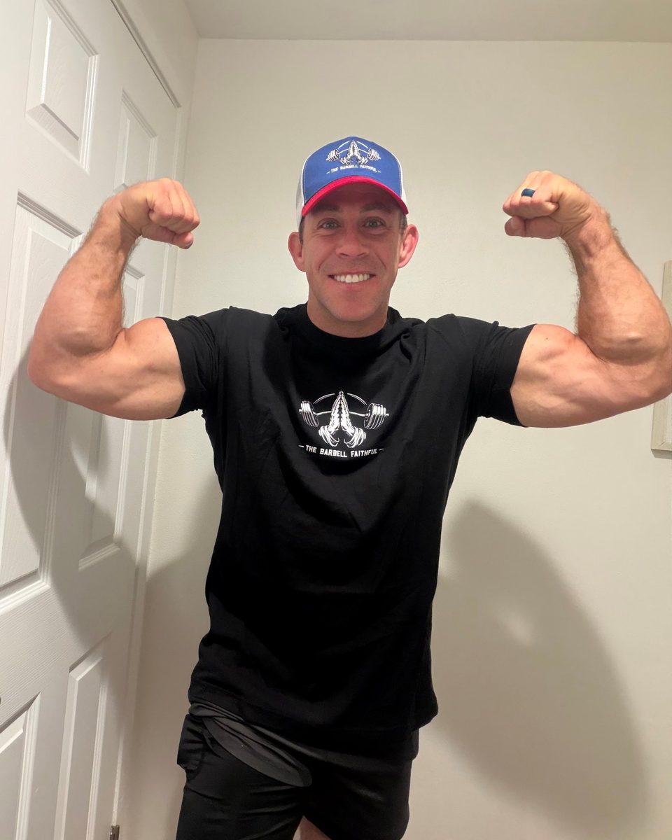 Mike flexing in the red, white and blue Barbell Faithful trucker hat and black tee